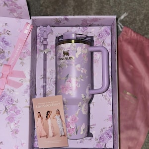 K&ouml;nnte beinhalten: Ein lavendelfarbener Stanley-Becher mit Blumenmuster, passendem Strohhalm mit Schleifenaufsatz und einer LoveShackFancy-Karte in einer floralen Geschenkbox. Der Becher verf&uuml;gt &uuml;ber einen Griff, einen Deckel und das schwarze Stanley-Logo.