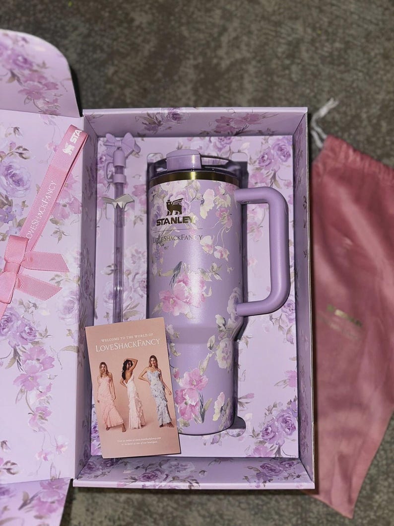 K&ouml;nnte beinhalten: Ein lavendelfarbener Stanley-Becher mit Blumenmuster, passendem Strohhalm mit Schleifenaufsatz und einer LoveShackFancy-Karte in einer floralen Geschenkbox. Der Becher verf&uuml;gt &uuml;ber einen Griff, einen Deckel und das schwarze Stanley-Logo.