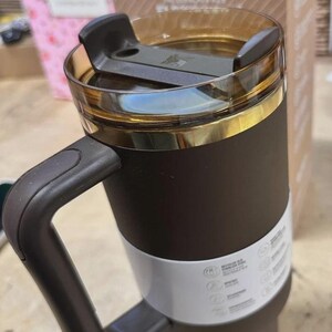 May include: A dark brown Stanley tumbler with a matching handle and a translucent amber-tinted lid. The cup features a white paper band around the center with printed text and a gold-colored rim accent. The tumbler is positioned in front of its box.
