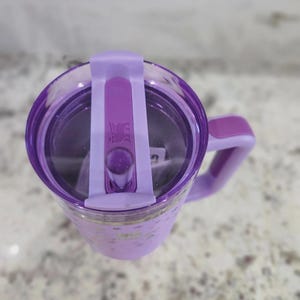 May include: A light purple tumbler with a matching handle and a clear lid featuring a purple sliding closure. The lid displays a small embossed bear logo. The tumbler body has a subtle star pattern and a smooth finish, viewed from directly above.
