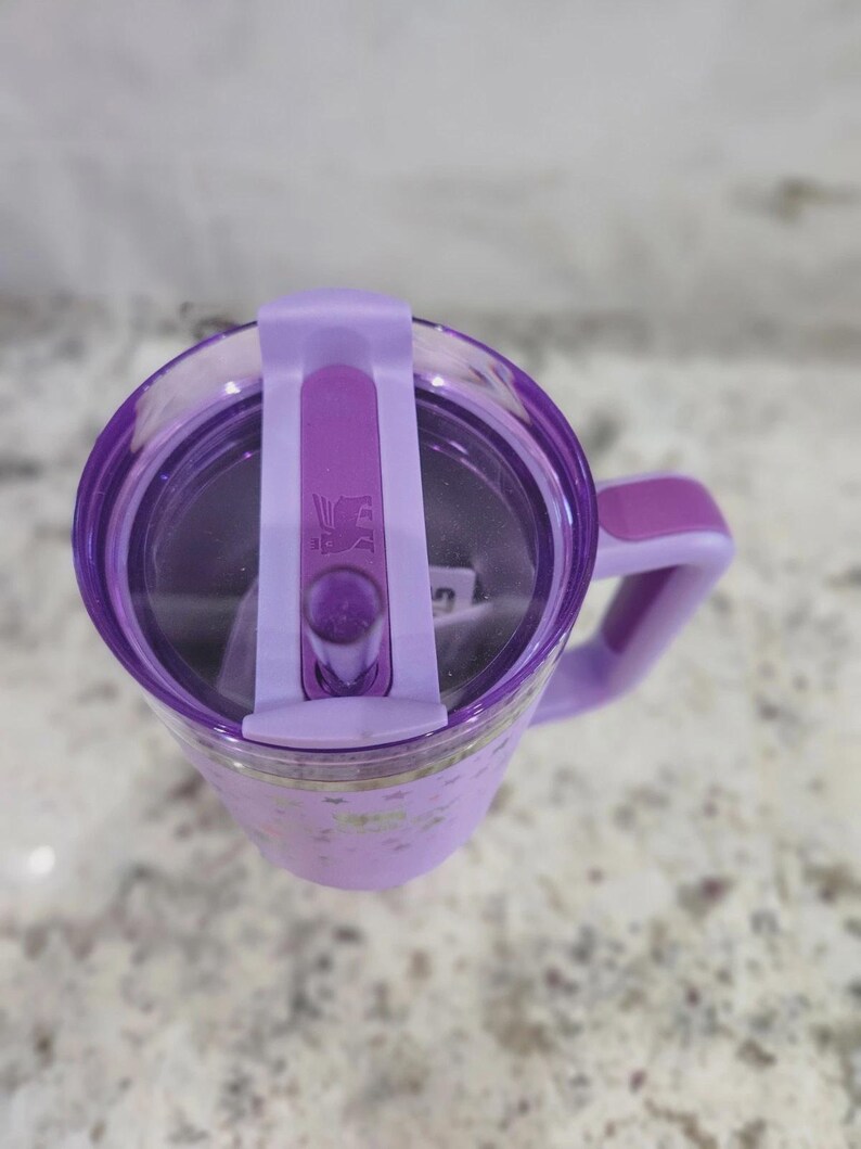 May include: A light purple tumbler with a matching handle and a clear lid featuring a purple sliding closure. The lid displays a small embossed bear logo. The tumbler body has a subtle star pattern and a smooth finish, viewed from directly above.