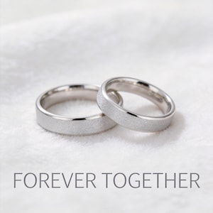 Puede incluir: Dos anillos de boda plateados con un acabado texturizado, sobre una superficie blanca. Los anillos están uno al lado del otro, con el texto "FOREVER TOGETHER" debajo.