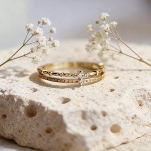 Puede incluir: Un anillo dorado con dos bandas, una con pequeños elementos decorativos y la otra con diminutas piedras brillantes. Una pequeña piedra de cuatro puntas está engastada en el centro. El anillo se exhibe sobre una superficie texturizada de color claro.