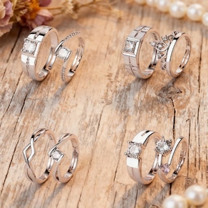 Puede incluir: Cuatro pares de anillos de plata con piedras preciosas transparentes se muestran sobre una superficie de madera. Los anillos tienen varios diseños, incluyendo una cruz, un patrón geométrico y una banda delicada con pequeñas piedras.