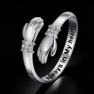 Könnte beinhalten: Ein silberner Ring mit zwei sich umarmenden Händen, verziert mit funkelnden Details. Der Ring ist mit "Always in My heart" graviert. Das Bild zeigt den Ring vor schwarzem Hintergrund.
