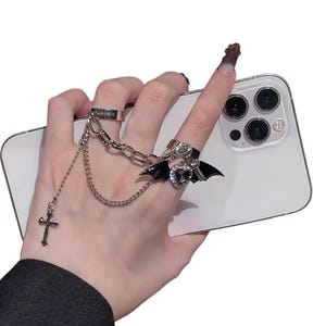 May include: A silver-toned ring set featuring a chain and cross pendant. The set includes a ring with the word "simple" engraved, and a ring with a black bat wing design and a heart-shaped gem. The rings are connected by a chain.