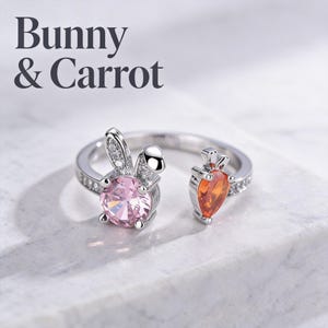 May include: A silver ring with a bunny and carrot design. The bunny features a pink gemstone and the carrot has an orange gemstone. The ring is set against a white background. The text "Bunny & Carrot" is at the top.