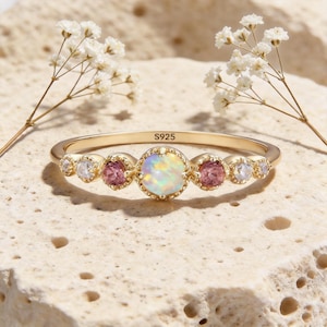 May include: A gold ring featuring an opal centre stone, with pink and clear gemstones either side. The ring is set on a textured surface with the text "S925". Delicate white flowers are in the background.