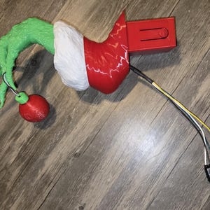 May include: A whimsical Christmas-themed decoration featuring a green hand holding a red ornament. The hand emerges from a red and white sleeve, connected to a red rectangular component with wires and a connector. The overall design evokes a festive, playful aesthetic.