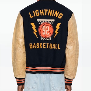 May include: A navy blue and tan varsity jacket with the words "LIGHTNING BASKETBALL" and a basketball graphic on the back. The jacket has tan leather sleeves and orange and black striped trim.