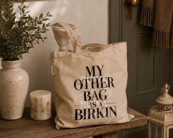 May include: A beige canvas tote bag featuring the black printed text MY OTHER BAG IS A BIRKIN. The bag has two fabric handles and a small rectangular tag on the side. The material appears to be a natural cotton fabric with a visible weave texture.
