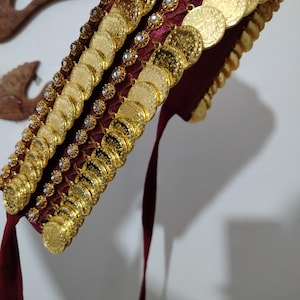 Può includere: Un copricapo decorativo con file di monete color oro e piccole gemme trasparenti. Il copricapo è attaccato a una base in tessuto bordeaux con nastri bordeaux. Le monete sono circolari e sembrano essere in rilievo.