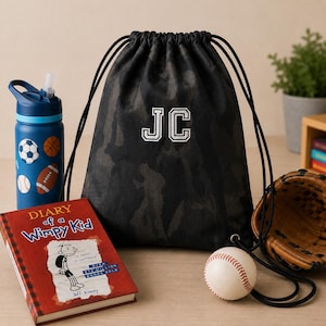 May include: A black camouflage drawstring bag featuring the embroidered white letters JC, a blue water bottle with sports ball graphics, a red Diary of a Wimpy Kid book, a baseball, and a brown leather baseball glove arranged on a flat surface.