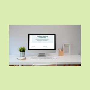 May include: A modern computer setup on a white desk. The screen displays "Keyboard Shortcuts for Beginners." A keyboard, mouse, small plant, notepad, pencils, and a picture frame are also on the desk. The background is a light gray wall.