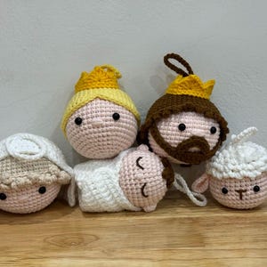 May include: A collection of handmade crochet Christmas ornaments. The set includes a baby, two kings with crowns, and two sheep. The ornaments are primarily pink, white, yellow, and brown, with black eyes. They are displayed on a wooden surface.
