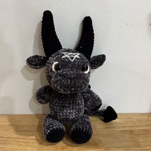 Może przedstawiać: Pluszowa, szara i czarna szydełkowa koza z dużymi czarnymi rogami, czarnym ogonem i białym pentagramem na czole. Maskotka siedzi na drewnianej powierzchni na tle białej ściany.