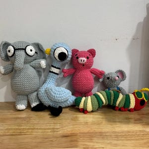 May include: A collection of handmade crochet stuffed animals, including an elephant wearing glasses, a blue pigeon, a pink pig, a gray mouse, and a colorful caterpillar. The toys are arranged on a wooden surface.