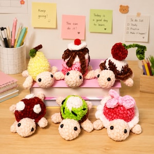 May include: Six plush crochet turtles with shells shaped like various foods, including a yellow pear, a pink dessert with chocolate drizzle and a cherry, a brown sundae, a red berry, a green kiwi, and a pink frosted treat with a bow.