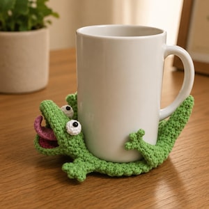 May include: A white ceramic mug rests on a green crocheted alligator coaster. The coaster features white eyes, a pink tongue, and is made of green yarn. The mug and coaster are on a wooden surface.