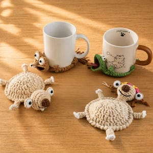 May include: Handmade crochet coasters in the shape of cartoon creatures, including a frog and a dog, with two white ceramic mugs. One mug has the text "I JUST WANT A". The coasters are in various shades of brown and beige.