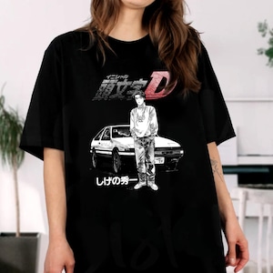 May include: Black t-shirt featuring a graphic of a white car and a person in a white shirt and jeans. Japanese text is above and below the image. The graphic is in black and white.