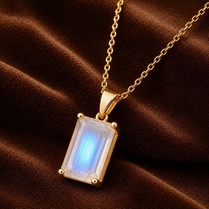 May include: A gold necklace featuring a rectangular emerald-cut moonstone pendant. The stone displays a translucent appearance with a central blue adularescence. The pendant is held by four prongs on a gold setting attached to a delicate gold chain.