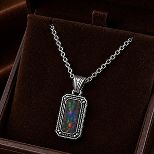May include: A silver-toned rectangular pendant necklace featuring a dark inlay with iridescent flecks of green, blue, and purple. The pendant is suspended from a silver-toned chain and is presented inside a brown velvet-lined jewelry box.
