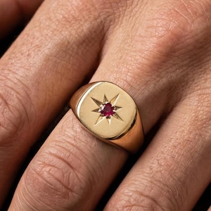 Puede incluir: Un anillo de sello dorado con un diseño de estrella y una pequeña gema roja en el centro. El anillo se lleva en un dedo, mostrando su acabado pulido y su estilo clásico.