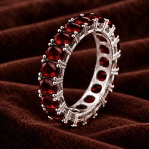 Op de afbeelding: Een zilveren eternity-ring met een doorlopende rij ovaal geslepen, dieprode granaat-edelstenen in een klauwzetting. De metalen band heeft een gepolijste afwerking en de stenen zijn rondom de gehele omtrek van de ring geplaatst.