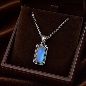 Könnte beinhalten: Eine rechteckige Anhängerkette mit einem zentralen, blau schimmernden Stein in einer silberfarbenen Metallfassung mit dekorativen Perlen- und Kordelverzierungen, hängend an einer passenden silberfarbenen Gliederkette.