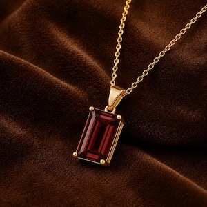 May include: A gold necklace featuring a rectangular emerald-cut deep red gemstone pendant held by four prongs. The pendant is attached to a delicate gold chain, showcasing a polished metal finish and a faceted stone surface with a rich crimson hue.