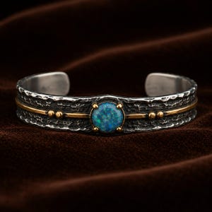 May include: A cuff bracelet featuring a dark, hammered silver band with a central circular blue opal stone set in gold prongs. A thin gold horizontal line runs across the center of the band, accented by small gold spheres on either side of the stone.
