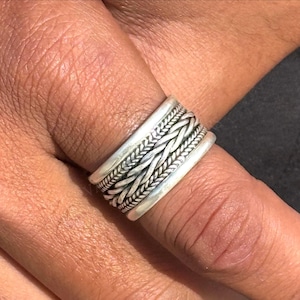 Peut inclure: Une bague en argent avec un motif tressé. La bague présente plusieurs bandes avec un motif texturé et tissé. La bague est portée sur un doigt, mettant en valeur les détails complexes du bijou.