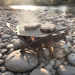 May include: A portable, hexagonal metal grill with integrated handles, positioned over glowing embers. Two round meat patties are cooking on the slatted surface, with visible steam rising from the heat source and a sturdy, angular frame design.