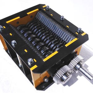 Può includere: Un trituratore industriale con un telaio nero e giallo. Il trituratore è dotato di più lame rotanti e ingranaggi, progettati per l'elaborazione dei materiali. L'immagine mostra il meccanismo interno e gli ingranaggi esterni.