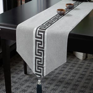 Pode incluir: Um caminho de mesa cinza com um desenho de chave grega preta no centro e uma borda prateada. O caminho de mesa tem uma borla azul escura na extremidade. Duas pequenas tigelas marrons estão sobre o caminho de mesa. O caminho de mesa está sobre uma mesa preta.