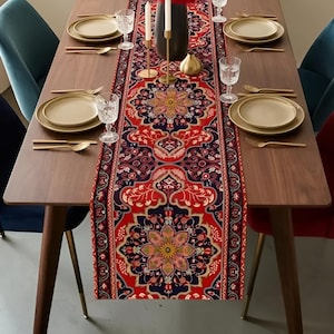 May include: A wooden dining table set with a decorative runner. The runner features a red, navy, and gold floral pattern. Gold-coloured plates, silverware, and clear glasses are set for dining. Teal and red chairs surround the table.