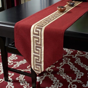 May include: A burgundy table runner with a gold and brown geometric pattern down the centre. The runner is draped over a black table, with two small wooden bowls on top. A tassel hangs from the end.