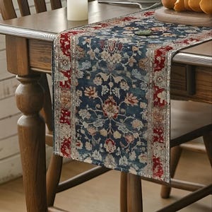 May include: A navy blue table runner with a floral pattern in red, cream, and light blue. The runner has a distressed, vintage look and is placed on a wooden table. The edges are trimmed with a red border.