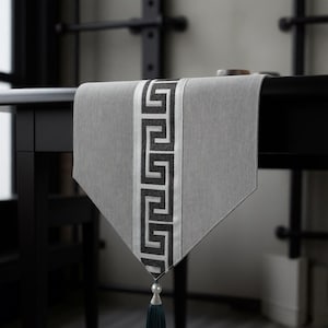 Puede incluir: Camino de mesa gris con un diseño de borde geométrico en blanco y negro. El camino de mesa tiene una borla con una perla y una borla de color verde azulado oscuro. El camino de mesa está sobre una mesa negra.