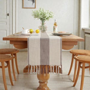 Puede incluir: Una mesa de comedor de madera con un camino de mesa a rayas beige y marrón. El camino de mesa tiene flecos en la parte inferior. La mesa está decorada con un jarrón de flores blancas, una botella y un plato. Tres taburetes de madera en primer plano.
