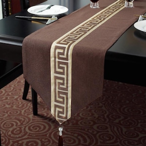 Op de afbeelding: Een bruine tafelloper met een centraal beige geometrisch ontwerp. De loper heeft een kwast aan het uiteinde en is op een zwarte tafel geplaatst. De loper is gemaakt van een textuurstof.