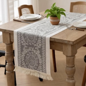 Puede incluir: Un camino de mesa con estampado gris y blanco con borde de flecos sobre una mesa de madera. Una pequeña planta en maceta se encuentra en el centro. Platos blancos y cubiertos están colocados sobre la mesa, con sillas en el fondo.