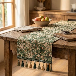 May include: A green floral table runner with a white and peach flower pattern. The runner has a scalloped edge and tassels in shades of green and beige. The runner is on a rustic wooden table.