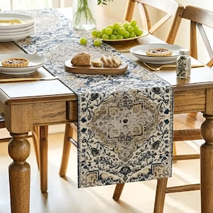 May include: A dining table set with a patterned table runner, plates, and food. The runner has an ornate design in blue, beige, and brown. Grapes and bread are on the table, creating a welcoming dining scene. The table is made of wood.