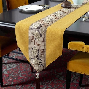 Peut inclure: Chemin de table jaune moutarde avec un panneau central à motifs floraux et géométriques beiges et marron. Une gland pend à l'extrémité. Le chemin de table est posé sur une table sombre, avec des chaises jaunes et un tapis rouge à motifs en arrière-plan.
