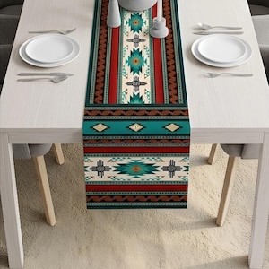 Puede incluir: Un camino de mesa rectangular con un diseño del suroeste. El corredor presenta un patrón repetitivo de formas geométricas en colores turquesa, rojo, marrón y crema. Se coloca sobre una mesa blanca, con platos y cubiertos blancos.