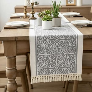 以下が含まれることがあります： 幾何学模様とフリンジトリムが付いた、グレーと白の模様のテーブルランナー。 ランナーは木製のテーブルの上に置かれ、お皿、カトラリー、鉢植えの植物が見えます。