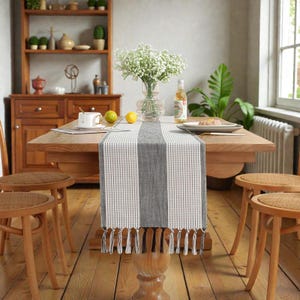 Puede incluir: Un camino de mesa a rayas grises y blancas con flecos, colocado sobre una mesa de madera. La mesa está decorada con un jarrón de flores blancas, un plato y una botella. La habitación cuenta con sillas de madera y un aparador.
