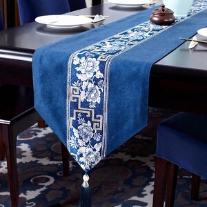 Könnte beinhalten: Blauer Tischläufer mit einem Blumendesign in Weiß und Gold. Der Läufer hat eine Quaste am Ende und liegt auf einem dunklen Holztisch. Der Läufer ist etwa 152 cm lang.